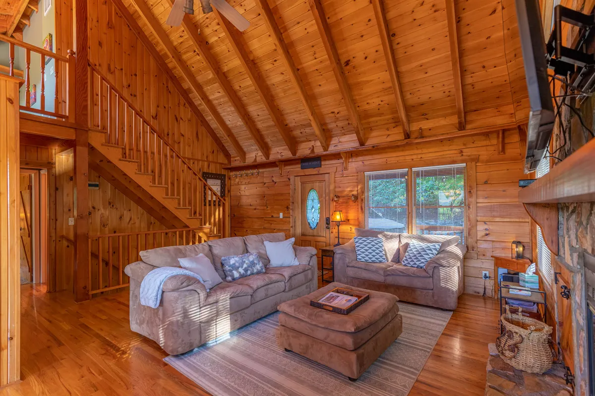 A welcoming living room scene with a comfortable sofa, coffee table, stone fireplace, wooden staircase, and ample windows letting in natural light. The cozy seating area invites guests to relax, socialize, or enjoy quiet moments by the fire.
