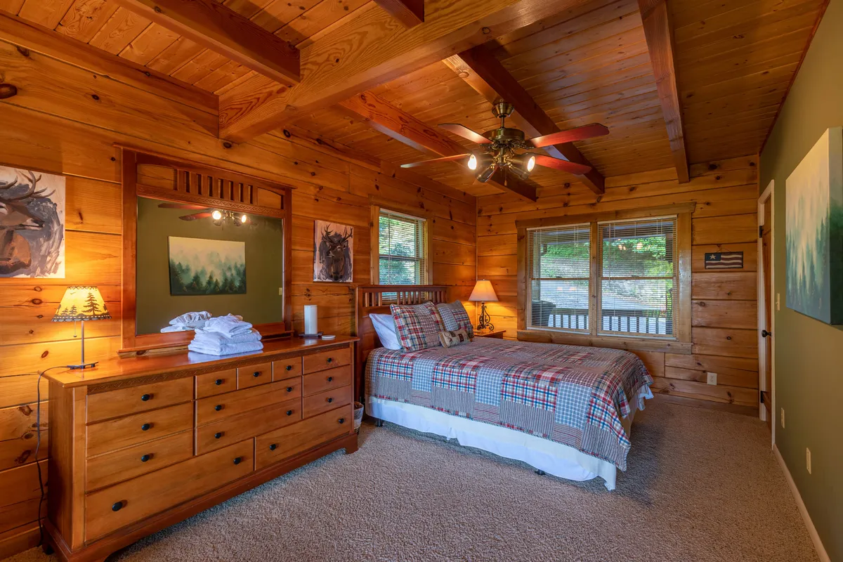 A cozy bedroom featuring a wooden bed, a dresser with a lamp, artwork on the wall, and large windows that allow natural light to flood the space. The relaxed atmosphere invites restful nights and quiet moments, perfect for unwinding after a day of activities.