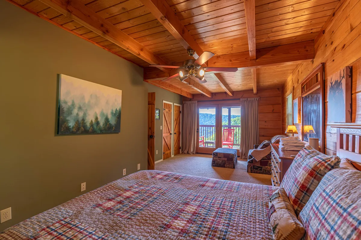 A warmly lit bedroom with a bed draped in a plaid quilt, wooden ceiling beams, and a large window offering scenic mountain views. The tranquil décor and intimate layout highlight the feeling of a personalized escape.