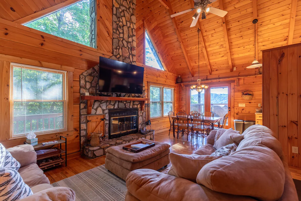 This image showcases a spacious living room featuring a striking stone fireplace, large windows flooding the area with natural light, a dining area, and comfortable seating. The vaulted ceilings and open floor plan are visually prominent, emphasizing the sense of openness and connection between living and dining spaces.