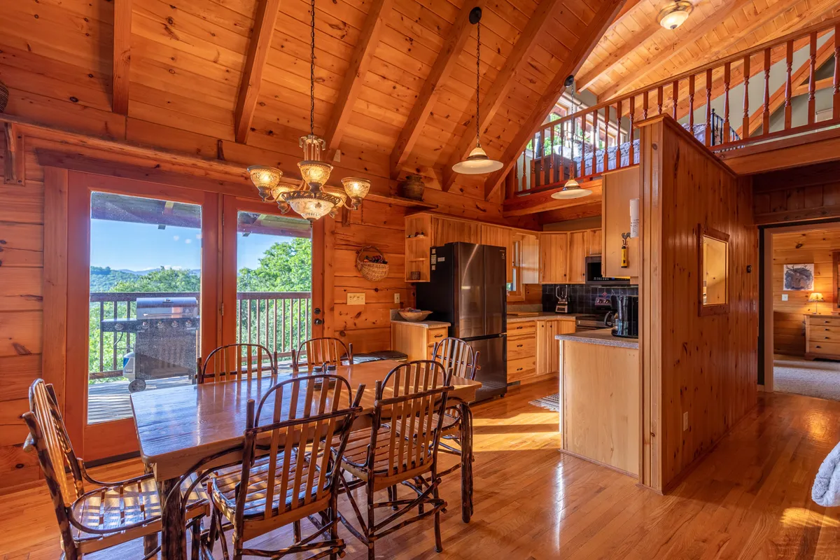 The photo highlights an open-concept kitchen and dining space with a wooden dining table, central kitchen island, and a striking chandelier. Large windows and a loft area add to the airy, inviting ambiance, reinforcing the seamless flow mentioned in the text.