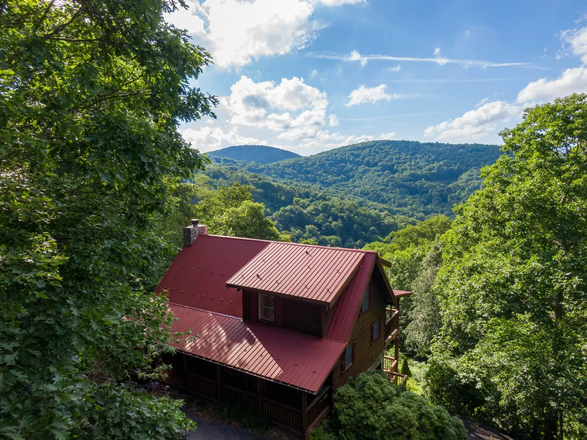 A charming wooden cabin with a distinctive red metal roof sits nestled among trees, with a sweeping mountain range visible in the background. The scene radiates calm and adventure, with the natural setting inviting guests to explore nearby trails or relax on the porch to take in stunning mountain vistas.