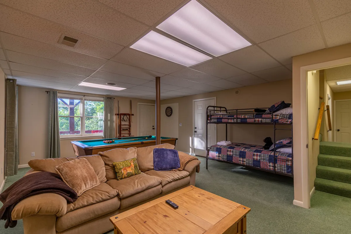 A wide interior shot reveals a relaxed basement lounge area with a sofa, twin bunk beds, pool table, and windows allowing in natural light. The arrangement communicates a multi-functional space ideal for both sleeping and socializing, echoing the description of the room as adaptable for various group needs.