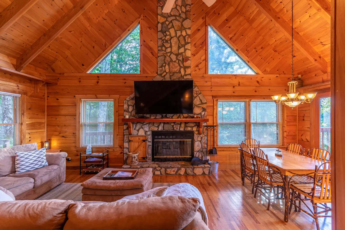 A warm, spacious interior featuring a striking stone fireplace, a large dining table, and expansive windows letting in natural light. Wooden beams and inviting decor make it clear this is a comfortable space for hosting dinners, game nights, or group celebrations.
