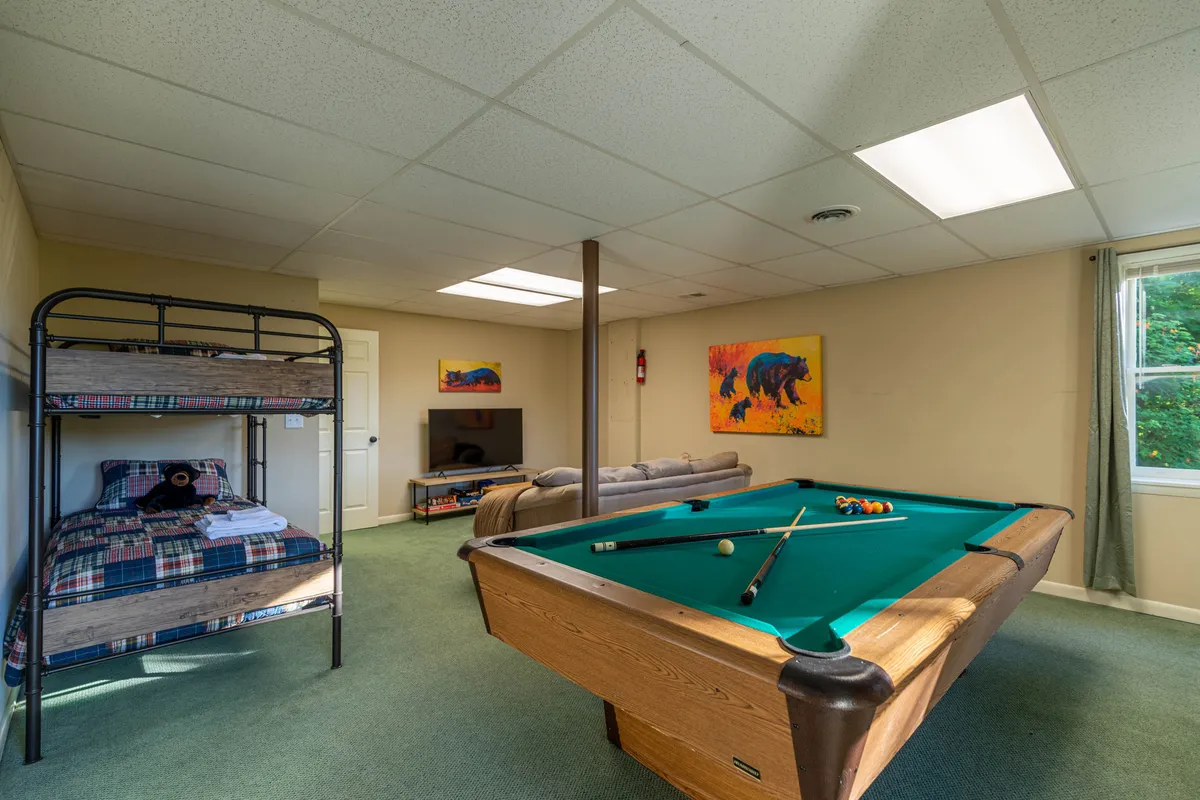 A playful basement area featuring a pool table, a twin bunk bed, a sleeper sofa, and a television. The space is decorated with artwork and offers ample room for games and relaxation, making it perfect for kids, teens, or extra guests.